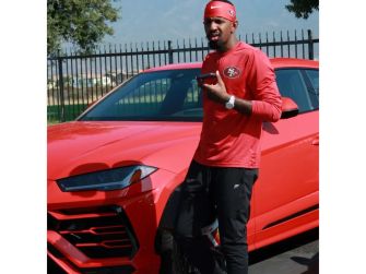 Daejon Love, wearing a red long-sleeve shirt and red headband, stands beside a red and a blue sports car in a parking lot, holding a San Francisco 49ers football helmet.
