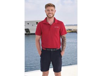 Nathan Gallagher stands at a marina by the water, wearing a red polo shirt and dark shorts, smiling toward the camera.