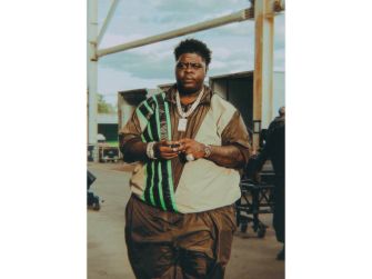 Xavier Landum, also known as BigXthaPlug, stands outdoors in a tan and green outfit with a gold chain, hands clasped in front.