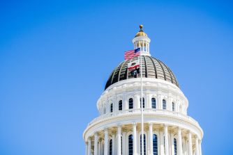 Dome of the California State Capitol rising against a clear blue sky with flags flying from flagpoles around the structure.