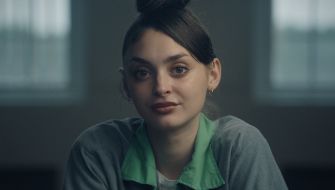 Mackenzie Shirilla sits indoors with a neutral expression, wearing a green-collared shirt under a gray sweater and facing the camera.