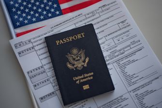 A United States passport rests on a government form with MOTHER and FATHER fields visible, and a U.S. flag in the background.