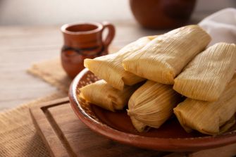 Tamales wrapped in corn husks piled on a brown ceramic plate on a wooden table, with a mug visible in the background.
