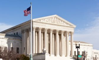The U.S. Supreme Court building with tall marble columns and a flag flying outside against a clear blue sky.