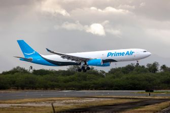 Prime Air Boeing 767 cargo jet on final approach to a runway, gear down, with a grassy area and trees visible in the background.