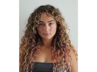 Gabbie Gonzalez with long curly hair, wearing a dark top, gazes straight at the camera.