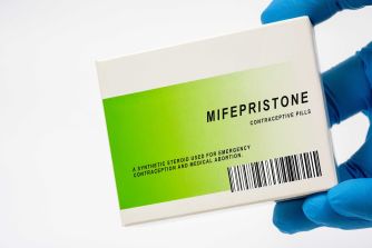 Box labeled MIFEPRISTONE with a green stripe and barcode is being held by a person wearing blue medical gloves.