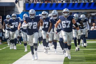Dallas Cowboys football players in navy uniforms and helmets walk onto the field.