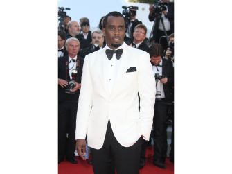 Sean “Diddy” Combs in a white tuxedo jacket and bow tie on a red carpet surrounded by photographers.