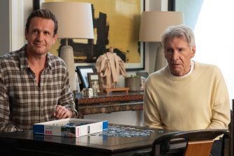 Jason Segel and Harrison Ford sit at a table with a board game in a living room.