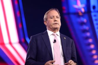 Texas Attorney General Ken Paxton speaks onstage in a dark suit and pink tie.