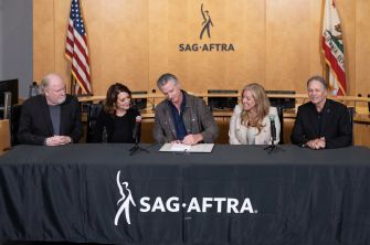 Five people sit behind a SAG-AFTRA table as a man in the center signs a document, with microphones, flags and the SAG-AFTRA logo visible.