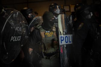Law enforcement officers in tactical gear stand together, holding shields marked “ICE Police.”