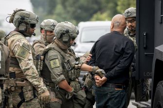 Several armed officers restrain a handcuffed man beside a vehicle.