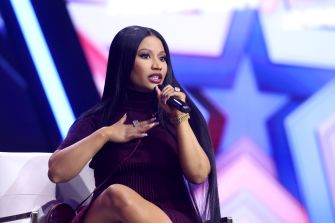 Nicki Minaj performs into a microphone onstage against a colorful star-shaped backdrop.