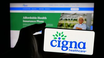 Cigna Healthcare logo on a card held in front of a laptop displaying an Affordable Health Insurance Plans webpage.