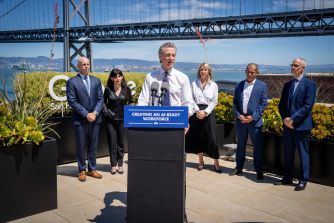 California Gov. Gavin Newsom speaks at a podium during an outdoor event with officials, with a bridge visible behind him.