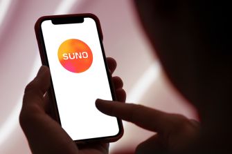 A person holds a smartphone displaying the Suno logo while another finger touches the screen.