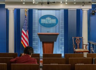 Empty White House briefing room with a podium, American flag, and a seated attendee.