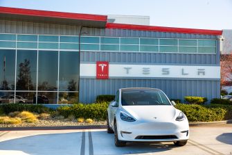 White Tesla parked outside a Tesla facility with the company logo visible on the building.