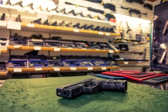 Handgun displayed on a green counter in a gun store, with rifles and other firearms visible on wall racks behind it.