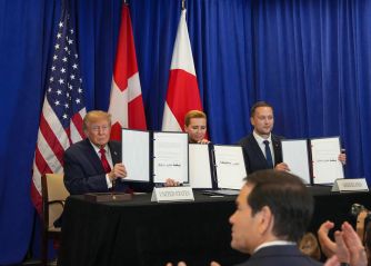President Donald Trump signs a security agreement alongside Danish Prime Minister Mette Frederiksen and Greenland Prime Minister Jens-Frederik Nielsen at a table in front of U.S., Danish and Greenlandic flags.