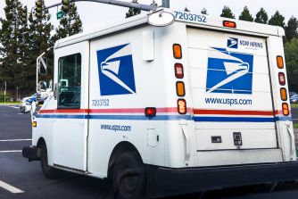 USPS mail truck parked in a lot, with the Postal Service logo and “Now Hiring” sign visible.