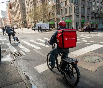 DoorDash delivery worker with a red insulated backpack rides an e-bike through a New York City intersection.