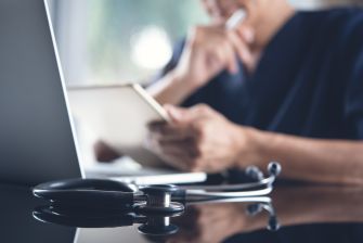 Healthcare professional holds a tablet beside a laptop and stethoscope.