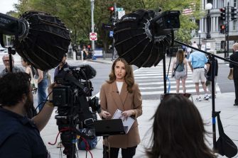 A female television journalist reports outdoors while camera operators and large studio lights surround her.