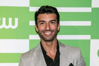 Justin Baldoni poses at an event in a gray blazer against a green-and-white backdrop.