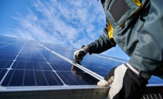 Solar panel worker in protective gear inspects panels on a rooftop under a partly cloudy sky.