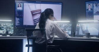 Woman in a control room examines computer monitors displaying identification photos and surveillance footage.