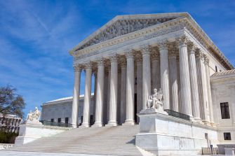 U.S. Supreme Court building with its columns, steps, and exterior statues under a blue sky.