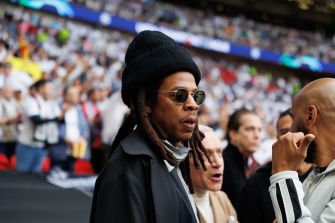 Jay-Z, wearing a black knit cap and sunglasses, speaks with a man in a stadium concourse.