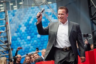 Arnold Schwarzenegger holds a microphone while standing onstage before a crowd.