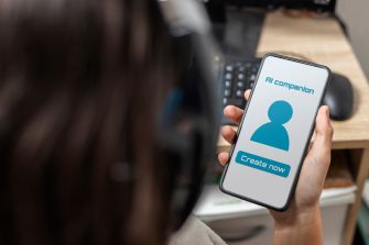Person holds a smartphone displaying an AI companion app’s profile icon and “Create now” button beside a laptop.