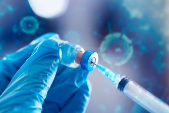 Gloved hands draw liquid from a vaccine vial into a syringe against a blue background of virus-like particles.