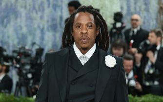 Jay-Z, also known as Shawn Carter, poses in a black suit with a white boutonniere as photographers gather behind him.