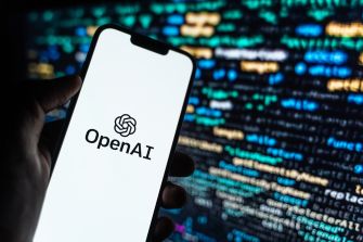 Hand holding a smartphone displaying the OpenAI logo against a background of computer code.