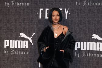 Rihanna poses in black attire at a Fenty fashion event with Puma and Fenty branding behind her.