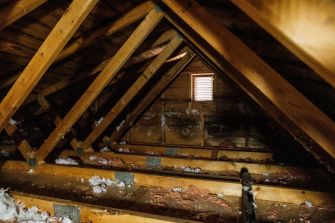 Unfinished attic with exposed wooden beams, insulation, debris, and a small window.