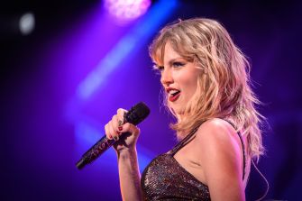 Taylor Swift sings into a microphone during a concert under purple and blue stage lights.