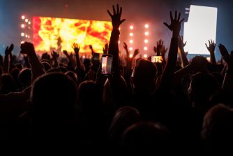 Concert crowd raising hands and recording with smartphones in front of a brightly lit stage.