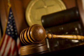 Judge’s gavel rests on a wooden sound block in a courtroom scene, with a U.S. flag and clock in the background.