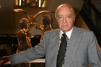 Mohamed Al-Fayed stands beside a bronze statue indoors, wearing a gray suit and extending one arm.