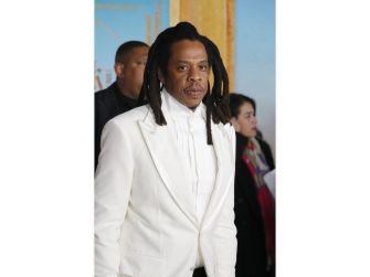 Jay-Z, also known as Shawn Carter, wearing a white suit and walking indoors.