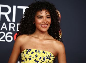 Olivia Dean smiles at a red-carpet event wearing a strapless yellow dress with blue floral patterns.