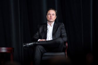 Elon Musk sits in a chair onstage against a black backdrop.