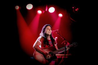 Gigi Perez performs on acoustic guitar beneath red stage lights.